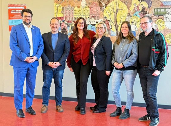 Sieben Personen stehen nebeneinander in einem Innenraum auf einem roten Boden vor einer hellen Wand. Die Gesichter sind unscharf dargestellt. Die Personen tragen unterschiedliche Kleidung, darunter Anzugjacken, Hemden, Jeans, Stoffhosen, Sneakers und Lederschuhe. Eine Person links trägt einen blauen Anzug, andere tragen dunkle Jacken oder Blazer, eine Person eine rote Bluse. Im Hintergrund hängt ein großes, farbiges Wandbild mit stilisierten Gebäuden, Bäumen und Menschen in einer Stadtszene. Links an der Wand befindet sich ein rotes Plakat mit weißer Schrift; der Text ist nicht vollständig lesbar.