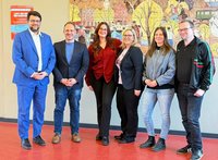 Sieben Personen stehen nebeneinander in einem Innenraum auf einem roten Boden vor einer hellen Wand. Die Gesichter sind unscharf dargestellt. Die Personen tragen unterschiedliche Kleidung, darunter Anzugjacken, Hemden, Jeans, Stoffhosen, Sneakers und Lederschuhe. Eine Person links trägt einen blauen Anzug, andere tragen dunkle Jacken oder Blazer, eine Person eine rote Bluse. Im Hintergrund hängt ein großes, farbiges Wandbild mit stilisierten Gebäuden, Bäumen und Menschen in einer Stadtszene. Links an der Wand befindet sich ein rotes Plakat mit weißer Schrift; der Text ist nicht vollständig lesbar.