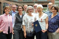 Mehrere erwachsene Personen stehen dicht nebeneinander im Freien vor einem Backsteingebäude. Eine Person in der vorderen Reihe hält eine dunkelblaue Urkundenmappe mit Bundesadler und trägt einen Orden an der Kleidung. Die Gruppe besteht aus neun Personen, die in zwei Reihen angeordnet sind. Der Hintergrund zeigt einen Innenhof mit rotem Backstein, Fenstern und etwas Dekoration. Das Foto wirkt wie eine Gruppenaufnahme bei einer offiziellen Ehrung.