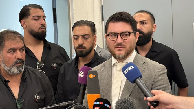 Eine Gruppe von Personen steht eng zusammen in einem Innenraum vor einer hellen Wand. Im Vordergrund sprechen mehrere Mikrofone mit sichtbaren Logos, darunter „ZDF“ und „RPR1“, in Richtung einer Person im grauen Anzug mit hellem Hemd und Krawatte sowie Brille. Um die sprechende Person herum stehen mehrere weitere Personen in dunkler Kleidung. Einige tragen runde Ansteckbuttons mit einem schwarz-weißen Porträtfoto. Hinter der Gruppe ist eine geöffnete Tür zu sehen.