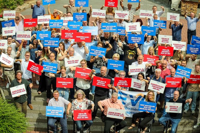 Große Gruppe von Menschen steht dicht beieinander im Freien und hält Schilder in die Kamera. Viele Schilder sind rot, blau oder weiß und tragen kurze Texte in Großbuchstaben sowie das Logo „EVG“. Einige Personen heben die Schilder über den Kopf, andere halten sie vor der Brust. Im Vordergrund sitzen einzelne Personen auf Stufen oder stehen auf einer Treppe; dahinter stehen weitere Menschen. Ein großes Banner mit der Aufschrift „EVG Eisenbahn- und Verkehrsgewerkschaft“ ist sichtbar. Die Umgebung zeigt Pflasterflächen, Treppen und etwas Grün im Hintergrund.