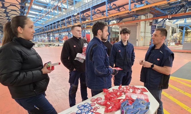 In einer großen Industriehalle stehen sechs Personen um einen rechteckigen Tisch. Der Tisch ist mit roten Stofftaschen, Aufklebern und kleinen Gegenständen bedeckt. Mehrere Personen tragen dunkle Arbeitskleidung, eine Person trägt eine schwarze Jacke. Eine Person hält einen kleinen Gegenstand in der Hand, eine andere einen Stapel roter Materialien. Im Hintergrund sind Hallenkonstruktionen aus Metall, Laufstege, Maschinen und eine weitläufige Produktionsfläche mit markierten Bodenlinien zu sehen.