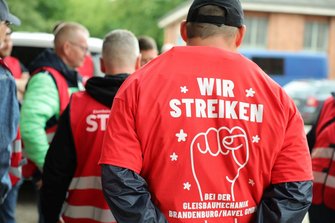 Rückansicht einer Person mit rotem T‑Shirt. Auf dem T‑Shirt steht in weißer Schrift: „WIR STREIKEN“ sowie darunter „BEI DER GLEISBAUMECHANIK BRANDENBURG/HAVEL GMBH“. Mehrere Personen in roten Westen stehen unscharf im Hintergrund.