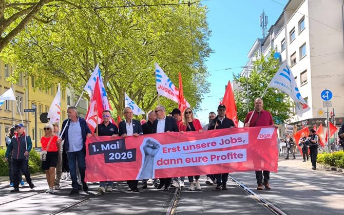 Eine Gruppe von Menschen läuft auf einer Straße mit Straßenbahnschienen. Mehrere Personen halten ein breites rotes Banner mit der Aufschrift „1. Mai 2026“ und „Erst unsere Jobs, dann eure Profite“, rechts daneben ein DGB-Logo und eine abgebildete erhobene Faust. Im Hintergrund sind Bäume, Wohngebäude und weitere Menschen mit Fahnen zu sehen, darunter rote Fahnen und weiße Fahnen mit blauen und roten Schriftzügen.