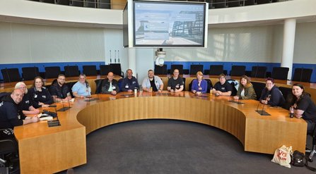 Großer Sitzungssaal mit halbkreisförmig angeordnetem Holztisch und integrierten Mikrofonen. Mehrere Personen sitzen nebeneinander am Tisch. An der Wand hängt ein großer Bildschirm mit einer Benutzeroberfläche; darüber verläuft eine Empore mit Geländer. Der Raum ist hell und weitläufig.