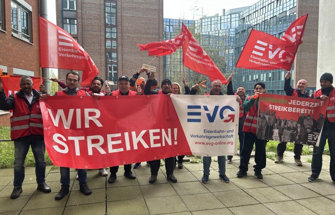 Eine Gruppe von Menschen steht im Freien auf einem gepflasterten Hof zwischen Backstein- und Glasgebäuden und hält mehrere große rote und weiße Fahnen und Banner. Auf einem roten Banner steht in weißer Schrift „WIR STREIKEN!“. Ein weißes Banner zeigt das Logo „EVG“ sowie den Text „Eisenbahn- und Verkehrsgewerkschaft“ und „www.evg-online.org“. Mehrere rote Fahnen tragen ebenfalls das EVG-Logo und den Schriftzug „Eisenbahn- und Verkehrsgewerkschaft“. Rechts ist ein weiteres rotes Banner mit der Aufschrift „JEDERZEIT STREIKBEREIT!“ zu sehen. Die Personen tragen überwiegend Jacken und Westen; im Hintergrund sind moderne Bürogebäude mit vielen Fenstern und etwas Grün zu erkennen.
