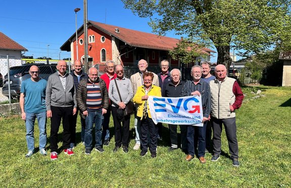 Gruppe von etwa vierzehn Personen, die im Freien auf einer grünen Wiese nebeneinander stehen. In der Mitte halten mehrere Personen ein blau-weißes Banner mit der Aufschrift „EVG Eisenbahn- und Verkehrsgewerkschaft“. Die Personen tragen unterschiedliche Freizeitkleidung wie Jacken, Westen, Pullover, Hemden, Jeans und Turnschuhe in verschiedenen Farben. Im Hintergrund steht ein rotes Gebäude mit Satteldach und hellen Fensterrahmen, daneben ein großer grüner Baum. Hinter der Gruppe sind ein Zaun, abgestellte Fahrzeuge und weitere Gebäude zu sehen. Der Himmel ist blau und das Bild ist bei Tageslicht aufgenommen.