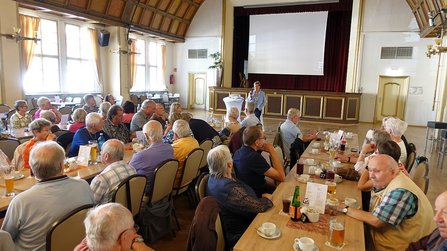 Großer Saal mit mehreren Tischen und zahlreichen sitzenden Personen. Im Vordergrund stehen zwei Personen vor einer Bühne mit rotem Vorhang und großer Projektionsleinwand. Auf der Leinwand ist eine Präsentation zu sehen. Hohe Fenster auf der linken Seite lassen Tageslicht in den Raum. Auf den Tischen stehen Getränke, Tassen und Tischaufsteller.