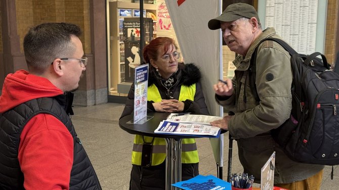 Drei Personen stehen in einer Bahnhofshalle an einem hohen Stehtisch mit Informationsmaterial. Auf dem Tisch liegen Flyer, Broschüren und ein Klemmbrett, daneben ein Behälter mit Stiften. Eine Person trägt eine gelbe Warnweste, eine andere eine rote Jacke, die dritte eine grüne Jacke mit Rucksack. Hinter dem Tisch steht ein aufgestelltes Banner, im Hintergrund sind Wände, Aushänge und weitere Passanten zu sehen.