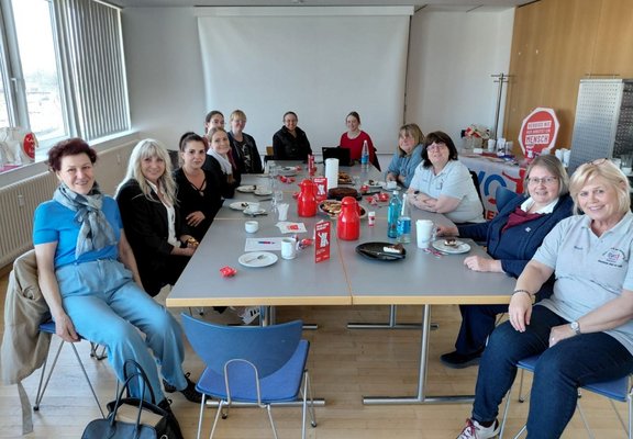 In einem hellen Besprechungsraum sitzen mehrere Personen um einen rechteckigen Tisch. Auf dem Tisch stehen Kaffeekannen in Rot, Tassen, Untertassen, Gläser, Wasserflaschen, Teller mit Gebäckresten und kleine rote Verpackungen. Die Personen sitzen auf Stühlen an allen Seiten des Tisches und blicken in Richtung Kamera. Im Hintergrund sind ein Fenster mit vertikalen Jalousien, eine Projektionsfläche sowie ein Roll‑up‑Banner und eine Fahne mit teilweise lesbarem Text zu sehen, darunter „Respekt“ und „Mensch“. Der Raum ist mit hellem Boden, schlichten Möbeln und Tageslicht ausgestattet.