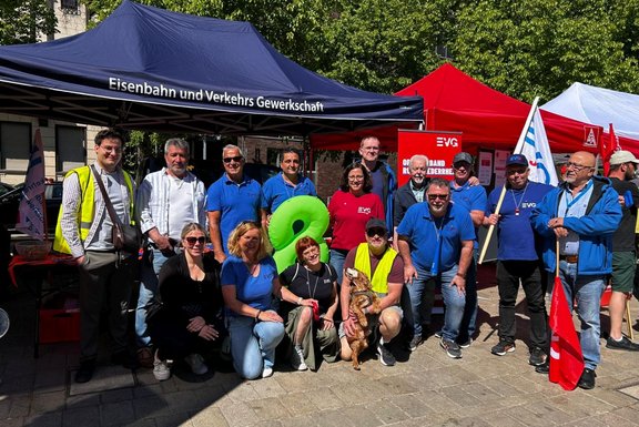 Gruppenfoto vor mehreren Pavillons im Freien. Ein blauer Pavillon trägt den Text „Eisenbahn und Verkehrs Gewerkschaft“. Daneben stehen rote und weiße Pavillons sowie ein rotes Banner mit der Aufschrift „EVG“ und „Ortsverband Ruhr-Niederrhein“. Mehrere Personen stehen und knien vor den Pavillons, eine Person hält eine Fahne. Im Vordergrund sitzt ein Hund auf dem Boden. Der Platz ist gepflastert, im Hintergrund sind Bäume und Gebäude sichtbar.