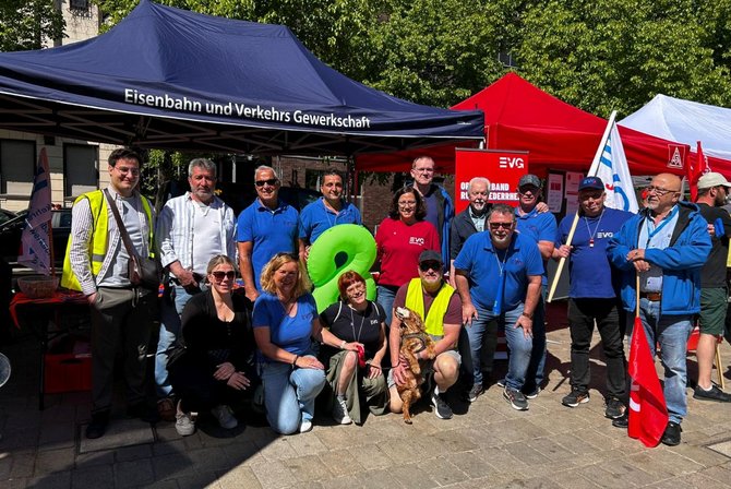 Gruppenfoto vor mehreren Pavillons im Freien. Ein blauer Pavillon trägt den Text „Eisenbahn und Verkehrs Gewerkschaft“. Daneben stehen rote und weiße Pavillons sowie ein rotes Banner mit der Aufschrift „EVG“ und „Ortsverband Ruhr-Niederrhein“. Mehrere Personen stehen und knien vor den Pavillons, eine Person hält eine Fahne. Im Vordergrund sitzt ein Hund auf dem Boden. Der Platz ist gepflastert, im Hintergrund sind Bäume und Gebäude sichtbar.