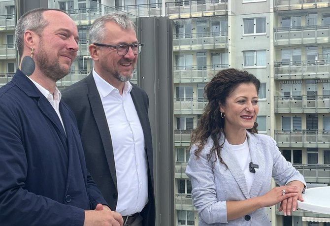 Drei Personen stehen auf einem Balkon oder einer Terrasse vor einer Fassade mit vielen Balkonen und Fenstern. Eine Person links trägt eine dunkle Jacke und einen großen dunklen Ohrring, die Person in der Mitte trägt einen dunklen Blazer über einem weißen Hemd, die Person rechts ein hell gestreiftes Sakko über einem weißen Oberteil. Im Hintergrund sind mehrstöckige Wohngebäude zu sehen.