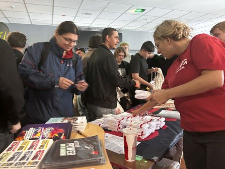 Informationsstand in einem Innenraum. Auf einem Tisch liegen Stapel weißer Textilien mit rotem Aufdruck, Schlüsselbänder, Flyer und Broschüren. Eine Person in rotem T-Shirt ordnet Material auf dem Tisch. Mehrere Personen stehen vor dem Stand und halten Taschen oder Informationsmaterial in den Händen. Auf dem Tisch steht ein Becher mit Deckel und Strohhalm. Im Hintergrund sind weitere Personen sowie Automaten und Wandflächen zu sehen.