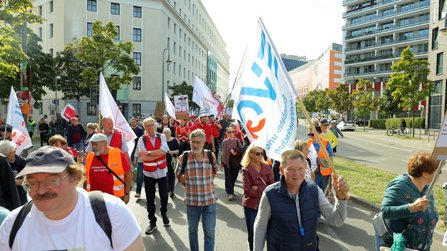 Menschen laufen auf einer städtischen Straße mit mehreren Fahnen und Transparenten. Mehrere Personen tragen orangefarbene Warnwesten. Im Hintergrund stehen mehrstöckige Gebäude, Bäume und weitere Teilnehmende des Demonstrationszuges.