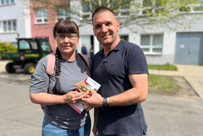 Zwei Personen stehen im Freien nah beieinander. Beide halten gemeinsam eine kleine rechteckige Karte sowie mehrere Papierflyer. Eine Person trägt ein graues T‑Shirt und einen Rucksack, die andere ein dunkles Kurzarmshirt und eine Armbanduhr. Im Hintergrund sind ein helles Gebäude mit Fenstern, ein Fahrzeug und weitere Personen auf einem Weg zu sehen.