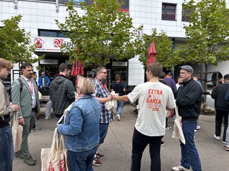 Außenbereich mit mehreren Personen auf einem Platz zwischen Bäumen und Sitzbänken. Viele Personen tragen helle Stofftaschen mit rotem Aufdruck. Im Hintergrund befinden sich Ladenfronten mit den Beschriftungen „Müller“, „SNACKS“ und „CAFÉ“. Eine Person im Zentrum trägt ein helles T-Shirt mit der Aufschrift „EXTRA GVNA GANZA NRW 2026“ auf dem Rücken. Mehrere Personen stehen in kleinen Gruppen und unterhalten sich.