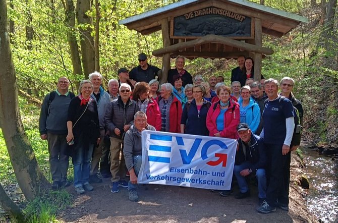 Eine Gruppe von etwa zwanzig Personen steht dicht beieinander auf einem Waldweg vor einem hölzernen Unterstand. Über dem Unterstand ist ein geschnitztes Schild mit der Aufschrift „Die Drachenschlucht“ und einer Drachenfigur angebracht. Die Personen tragen überwiegend Jacken und Outdoor-Kleidung. Im Vordergrund halten zwei Personen ein Banner mit der Aufschrift „EVG Eisenbahn- und Verkehrsgewerkschaft“. Umgeben ist die Szene von Bäumen, Laub und einem bewaldeten Hang.