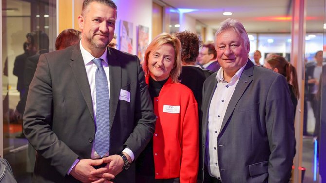 Drei Personen stehen nebeneinander in einem Innenraum. Alle tragen formelle Kleidung und Namensschilder. Die Person links trägt einen dunklen Anzug und eine gestreifte Krawatte. Die Person in der Mitte trägt eine rote Jacke über dunkler Kleidung. Die Person rechts trägt einen dunklen Anzug und ein helles Hemd. Im Hintergrund sind weitere Menschen sowie ein beleuchteter Flur mit Glaswänden zu sehen.