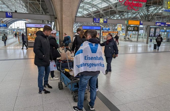 Große Bahnhofshalle mit Glasdach und hellgrauem Boden. Mehrere Personen stehen um einen kleinen Handwagen; eine Person trägt eine blau‑weiße Fahne mit dem sichtbaren Text „Eisenbahn‑ und Verkehrsgewerkschaft“. Weitere Personen gehen durch die Halle. Im Hintergrund sind Bahnsteig‑ und Wegeleitschilder mit den Nummern 13, 14 und 15, Anzeigetafeln, Geschäfte und Sitzbereiche zu sehen.