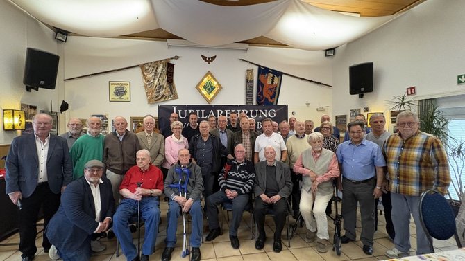 Große Gruppe von Personen in einem Saal, angeordnet in mehreren Reihen, einige sitzend auf Stühlen, andere stehend dahinter. Im Raum sind Fahnen, Wappen und dekorative Stoffbahnen an Decke und Wänden zu sehen. An der Rückwand hängt ein dunkles Banner mit der gut lesbaren Aufschrift „Jubilarehrung EVG Wupper Rhein Berg“. Links und rechts sind Lautsprecher an der Wand montiert. Der Boden ist gefliest, im Vordergrund stehen Stühle; einige Personen nutzen sichtbare Gehhilfen wie Rollatoren oder Gehstöcke.