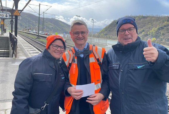 Drei Personen stehen auf einem Bahnsteig im Freien. Zwei tragen dunkle Jacken, eine Person in der Mitte zusätzlich eine orangefarbene Warnweste und hält ein weißes Blatt Papier in den Händen. Die Person rechts hebt den Daumen. Im Hintergrund sind Gleise, Oberleitungen, ein Bahnsteigschild, Geländer sowie eine hügelige Landschaft mit Bäumen und Himmel zu sehen.