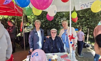 Drei Personen stehen hinter einem Infotisch unter einem weißen Pavillon mit bunten Luftballons. Auf dem Tisch liegen Süßigkeiten und Informationsmaterial. Ein rosa Luftballon mit „EVG“ ist sichtbar. Im Hintergrund sind weitere Personen, ein roter Pavillon und Bäume zu sehen.