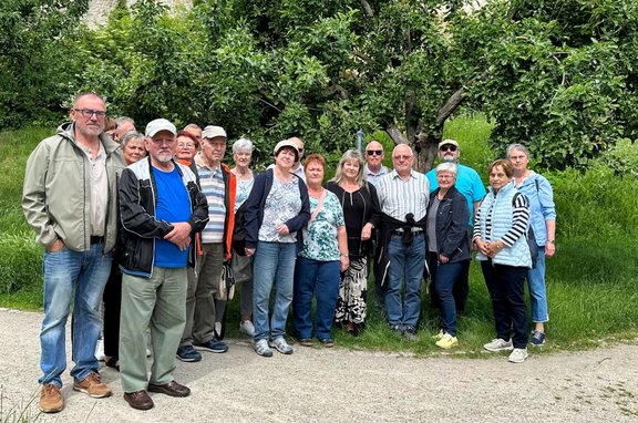 Gruppe von etwa 15 Personen steht auf einem Weg vor einer Wiese und mehreren Bäumen. Die Personen tragen überwiegend Freizeitkleidung, darunter Jacken, Hemden, Westen und Kappen. Im Hintergrund befinden sich dichtes Grün und Baumstämme; die Aufnahme entstand im Freien bei Tageslicht.