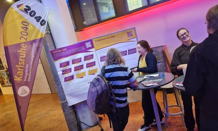 Ein Informationsstand in einem Innenraum mit einer Fahne „Karlsruhe 2040 Integriertes Stadtentwicklungskonzept“. Daneben stehen zwei große Schautafeln mit den Überschriften „Karlsruhe 2040“ und „Zielbild Karlsruhe 2040“. Mehrere Personen befinden sich an Stehtischen vor den Tafeln. An den Tafeln sind auch QR-Codes sichtbar.