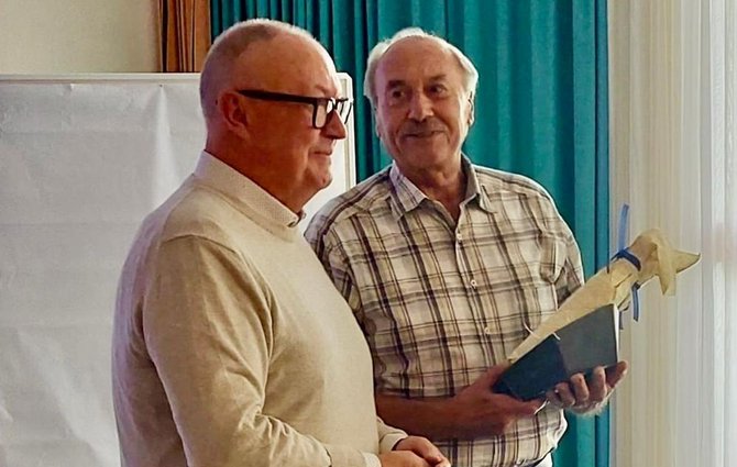 Zwei Personen stehen in einem Innenraum vor einem Flipchart und türkisfarbenen Vorhängen. Eine Person hält eine helle, sternförmige Holzskulptur auf einem dunklen Sockel in den Händen.