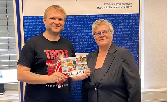 Zwei Personen stehen nebeneinander in einem Innenraum vor einer blauen Wandtafel. Beide halten gemeinsam einen rechteckigen Gutschein mit der Aufschrift „REISEGUTSCHEIN“, auf dem mehrere kleine Fotos von Landschaften und Freizeitmotiven abgebildet sind. Die Person links trägt ein schwarzes T‑Shirt mit rotem und weißem Aufdruck, die Person rechts trägt ein dunkles Jackett über einem dunklen Oberteil. Im Hintergrund ist auf der blauen Wandtafel viel weißer Text mit Berufsbezeichnungen zu sehen. Oben rechts an der Wand steht der Text: „EVG‑Geschäftsstelle Halle – Treffpunkt für unsere Mitglieder“. Links im Bild ist ein Fenster mit horizontalen Jalousien zu erkennen.