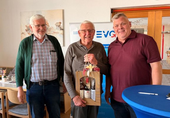 Drei Personen stehen nebeneinander in einem Innenraum. Die mittlere Person hält eine Holz-Geschenkverpackung mit zwei Flaschen und einer Schleife am Tragegriff. Die Personen tragen Freizeitkleidung: links eine grüne Strickjacke über einem karierten Hemd und Jeans, in der Mitte ein graues Langarmoberteil, rechts ein kurzärmeliges, dunkles Poloshirt. Im Hintergrund sind ein Roll-up-Banner mit teilweise sichtbarer Schrift, eine Glastür, Wanddekoration sowie Tische und Stühle zu sehen. Im Vordergrund rechts steht ein runder Stehtisch mit blauem Tischbezug und einem Stift darauf.