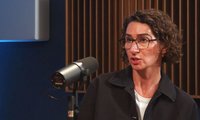 Person sitzt vor einem schwarzen Mikrofon auf einem Stativ. Der Gesichtsbereich ist von einem rechteckigen Farbfeld bedeckt. Die Person trägt eine dunkle Jacke über einem hellen Oberteil. Im Hintergrund sind senkrechte Holzleisten und eine dunkelblaue Wand zu sehen, links ein rechteckiges helles Element.