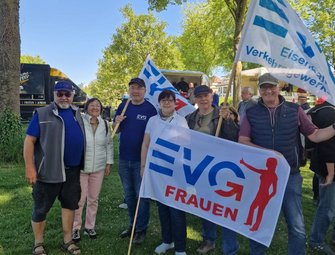 Gruppe von Personen steht im Freien auf einer Wiese mit Bäumen im Hintergrund. Mehrere Personen halten weiße Fahnen und Banner mit blau-roter Aufschrift. Auf einem Banner steht „EVG FRAUEN“, auf einer Fahne „EVG Eisenbahn- und Verkehrsgewerkschaft“. Im Hintergrund sind weitere Personen, ein Verkaufswagen und parkende Fahrzeuge zu sehen. Tageslicht, blauer Himmel.