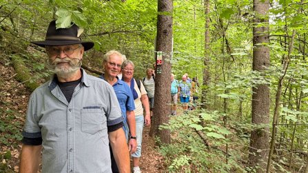 Eine Gruppe von Menschen geht in einer Reihe auf einem schmalen Waldweg. Im Vordergrund steht ein Mann mit Hut, Brille und auffälligem geschwungenem Schnurrbart. Hinter ihm folgen weitere Personen zwischen dicht stehenden Bäumen.