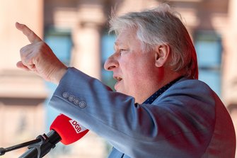 Seitenansicht einer Person an einem Mikrofon mit rotem Windschutz mit der Aufschrift „DGB“. Die Person trägt ein hellblaues Sakko. Ein Arm ist ausgestreckt, die Hand zeigt mit dem Zeigefinger nach vorne. Im Hintergrund sind helle Gebäudestrukturen und rote Lichtreflexe erkennbar.
