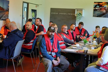 Mehrere Personen in roten Warnwesten sitzen an zusammengestellten Tischen in einem Besprechungsraum. Auf den Tischen stehen Tassen, Kaffeekannen, Blumenvasen und Unterlagen.