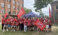 Gruppe von etwa 25 Personen auf einer Wiese vor Backsteingebäuden. Viele tragen rote Warnwesten. Mehrere rote Fahnen mit der Aufschrift „EVG“ sind zu sehen. Im Hintergrund steht ein blaues Zelt mit der Aufschrift „EVG Eisenbahn- und Verkehrsgewerkschaft“. Bäume und bewölkter Himmel im Hintergrund.