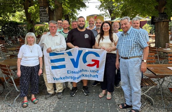 Eine größere Gruppe von Menschen steht im Außenbereich eines Biergartens unter Bäumen. In der Mitte halten mehrere Personen gemeinsam ein Banner mit der Aufschrift „EVG Eisenbahn- und Verkehrsgewerkschaft“. Um sie herum stehen weitere Personen, einige mit verschränkten Armen oder Händen vor dem Körper. Im Hintergrund sind Tische, Stühle und Schilder zu sehen.