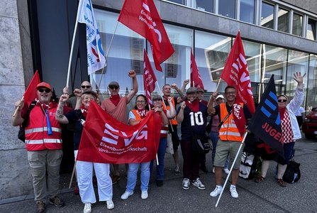 Gruppe von Personen steht vor einer Gebäudefassade und hält rote und schwarze Fahnen sowie Banner mit der Aufschrift „EVG“ und „EVG Jugend“. Einige tragen Westen in Signalfarben sowie rote Halstücher. Mehrere Personen haben die Arme erhoben. Im Hintergrund sind Fenster eines modernen Gebäudes zu sehen.