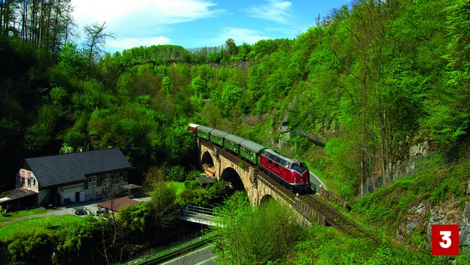 Rot-beige Lokomotive mit mehreren grünen Wagen auf einer steinernen Eisenbahnbrücke in einem bewaldeten Tal. Unter der Brücke verlaufen eine Straße und ein Bach. Die Hänge sind dicht mit grünen Bäumen und Sträuchern bewachsen. Unten rechts befindet sich eine rote Markierung mit der Zahl „3“.
