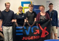 Fünf Personen stehen nebeneinander in einem Innenraum vor einer Wand und halten gemeinsam ein schwarzes Banner. Auf dem Banner befindet sich ein blau-rotes Logo mit der Aufschrift „EVG JUGEND“ sowie eine stilisierte rote Figur und ein Pfeil. Im Hintergrund sind an der Wand mehrere Tafeln und Plakate zu sehen, darunter ein großes Blatt mit Tabellen sowie ein blaues Schild mit der Aufschrift „Betriebsrat“. Die Personen tragen Freizeitkleidung, darunter T-Shirts, eine Bluse, Shorts und eine Umhängetasche. Einige tragen Brillen.