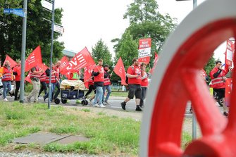 Demonstrationszug an einer Straßenkreuzung. Zahlreiche Personen tragen rote EVG-Fahnen und rote Westen oder T‑Shirts. Eine Person hält ein Schild mit der Überschrift „MERKT EUCH DAS:“. Im Vordergrund rechts ist ein unscharfes rotes Objekt zu sehen, links stehen Straßenschilder. Ein Bollerwagen mit EVG-Aufschrift wird mitgeführt.