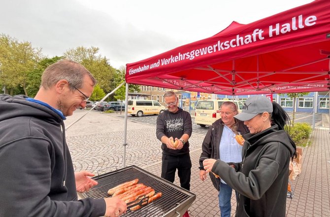 Mehrere Personen stehen im Freien unter einem roten Pavillon mit der Aufschrift „Eisenbahn und Verkehrsgewerkschaft Halle“. Im Vordergrund grillt eine Person auf einem rechteckigen Grill mehrere Bratwürste mit einer Grillzange. Andere Personen stehen um den Grill, eine Person hält ein Brötchen. Im Hintergrund sind gepflasterter Platz, parkende Fahrzeuge und Gebäude zu sehen.