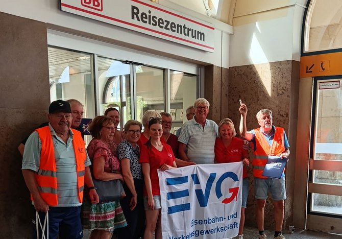 Gruppe von etwa zehn Personen steht in einem Innenraum vor einem Schalter mit der Aufschrift „DB Reisezentrum“. Mehrere Personen tragen orange Warnwesten, andere Alltagskleidung. In der Mitte halten sie ein weißes Banner mit der Aufschrift „EVG Eisenbahn- und Verkehrsgewerkschaft“. Eine Person rechts hebt eine Hand mit ausgestrecktem Zeigefinger. Hinter ihnen sind Glasflächen und ein Eingangsbereich sichtbar.