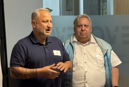 Zwei Personen stehen nebeneinander in einem Seminarraum vor einer Glaswand mit mattierten Streifen. Die Person links trägt ein dunkelblaues Poloshirt und gestikuliert mit den Händen. Die Person rechts trägt ein helles Hemd und eine hellblaue ärmellose Weste. Beide haben Namensschilder an der Kleidung. Im Hintergrund sind Fenster, Glasflächen und ein Pflanzenblatt sichtbar.
