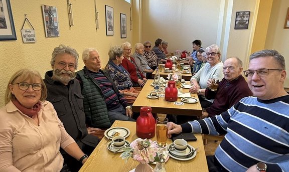Mehrere Personen sitzen in zwei Reihen an zusammengestellten Holztischen in einem schmalen Raum. Auf den Tischen stehen Kaffeetassen, Teller, Gläser und rote Thermoskannen sowie kleine Blumenarrangements. An den gelblichen Wänden hängen gerahmte Bilder und ein kleines Schild. Alle Personen sitzen dicht beieinander und blicken in Richtung Kamera.