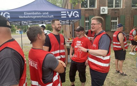 Mehrere Personen in roten Warnwesten stehen unter und vor einem blauen Pavillon mit der Aufschrift „EVG Eisenbahn- und Verkehrsgewerkschaft“. Im Hintergrund befinden sich ein Backsteingebäude, ein Tisch mit Getränken und weitere Personen. Auf einigen Kleidungsstücken sind die Aufschriften „EVG“, „STREIK!“ und „OHNE TARIF? OHNE MICH“ sichtbar.