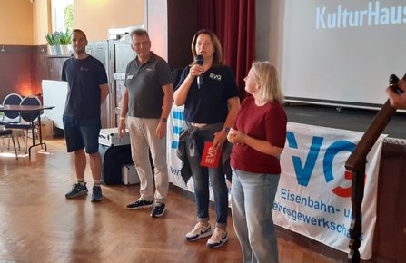 Innenraum mit kleiner Bühne vor einem roten Vorhang. Vier Personen stehen vor einem Banner mit der Aufschrift „EVG Eisenbahn- und Verkehrsgewerkschaft“. Eine Person hält ein Mikrofon. Im Hintergrund ist auf einer Projektionsfläche der teilweise sichtbare Schriftzug „KulturHaus“ zu sehen.