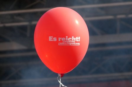 Roter Luftballon vor einem unscharfen Hintergrund aus Metallstreben. Auf dem Luftballon steht in weißer Schrift: „Es reicht! Das Leben bezahlbar machen.“ Am unteren Ende ist eine weiße Schnur befestigt.